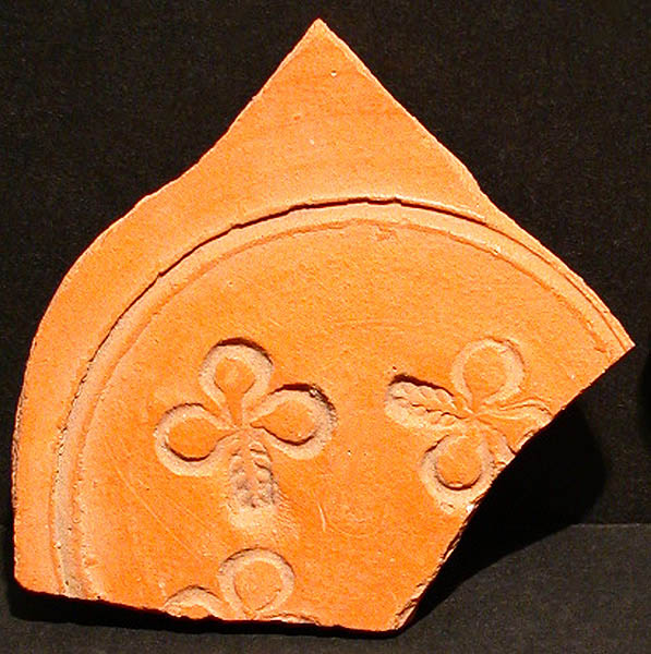 Fragment of a terra cotta vessel stamped with circle enclosing three repeating three-leaf clovers, two broken with some loss. Black background.
