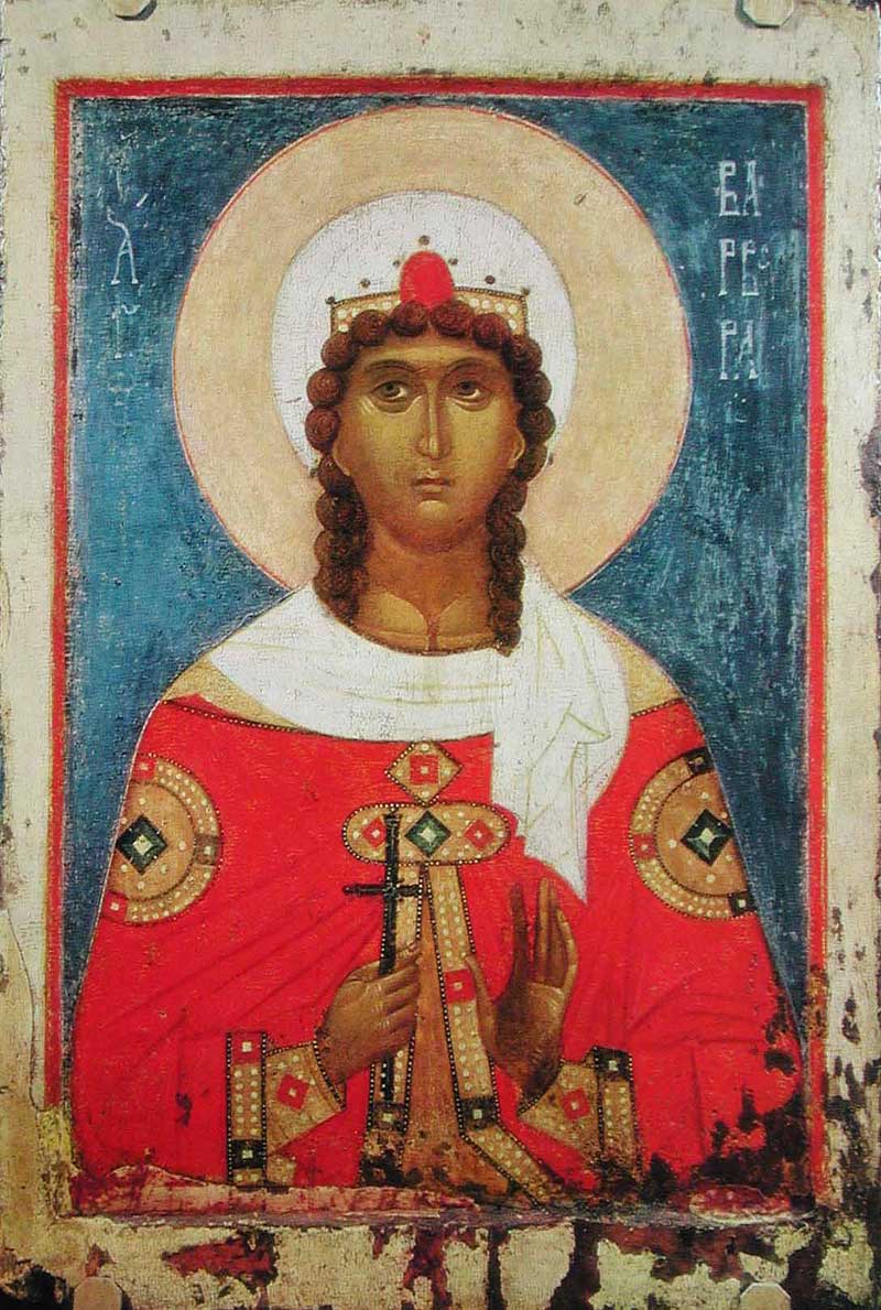 Half figure of woman with wavy brown hair, wearing jeweled crown and halo, and red and gold jeweled and pearl-trimmed garment, raising her open left hand and holding cross in her right hand. She is against a blue background with her name inscribed in white Greek letters H ΑΓΙA BAPBAPA, translated to English as Saint Barbara, and within red and white frame, with paint loss in some areas at bottom of the frame.