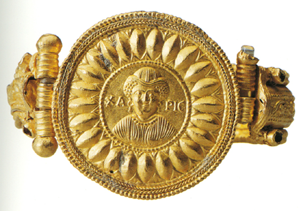 A gold bracelet with a large central medallion enclosing the bust of a woman, wearing a hat, earrings, and a necklace, inscribed in Greek with a word meaning Grace, Kindness, or Goodness. Hardware and hinges to attach the cuff on either side. Against a white background.