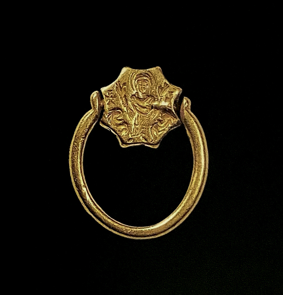 On the obverse of the rotating bezel of a gold ring, a woman with her hands raised stands between two animals and two crosses.