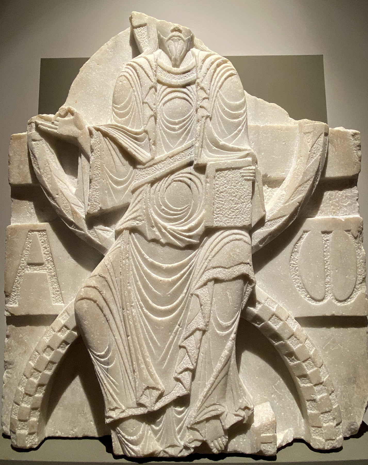 A fragment of white marble with a representation of a seated, bearded man with damaged head, holding a small round object in his right hand and a book in his left hand, flanked by two large letters. 