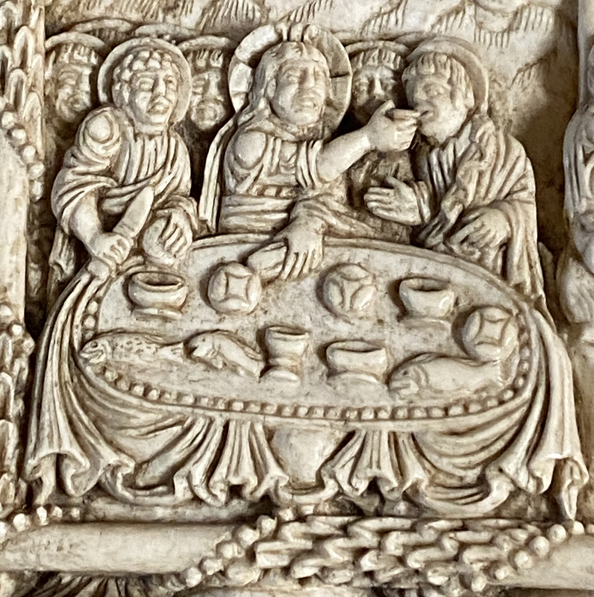A detail of an ivory plaque that contains a depiction of multiple men, of which three are in the foreground, behind a table containing fish, bread, bowls, and a chalice.