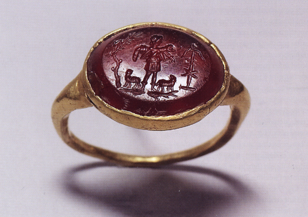Gold jewelry ring set with oval red gem carved with scene of a young man, wearing tunic, standing on fish, holding sheep over shoulders and flanked by two sheep and a tree with dove and anchor with monogram.
