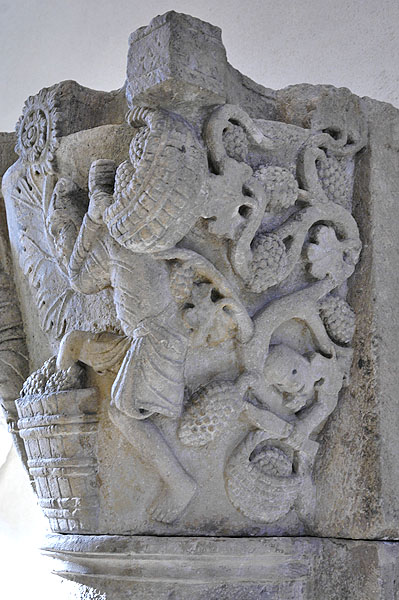 Detail of stone capital containing man carrying a basket of grapes on his shoulders and stepping into a vat of grapes with his left leg, in front of vines bearing grapes, with a man tasting a grape and holding basket full of grapes.