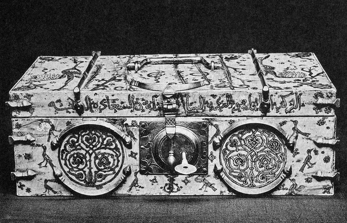 Front view of a rectangular ivory box with metal fittings, including a latch in front and a handle on the top. It is painted with animals, plants, and an Arabic inscription.