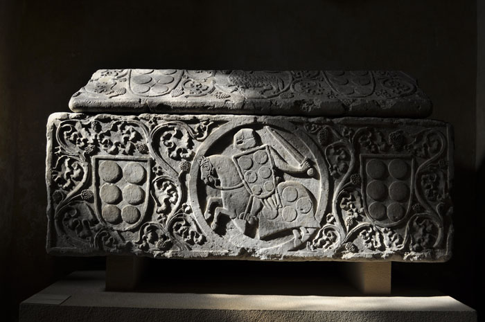 Tomb in shadows decorated with scrolling vines and shields and central medallion enclosing helmeted soldier holding sword and decorated shield, and mounted on draped horse.