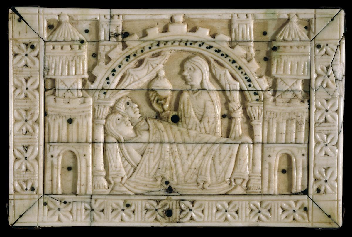 Rectangular carved ivory lid decorated with man and woman in bed with a woman holding covered vessel behind the bed, all in architectural niche with towers and rose ornament; cracks and drill holes.
