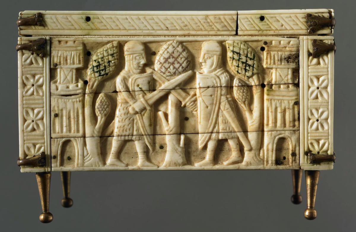 Rectangular panel of carved ivory casket depicting battling knights, holding shields, wielding swords, between trees and crenellated towers, rose ornament in border; cracks and drill holes.