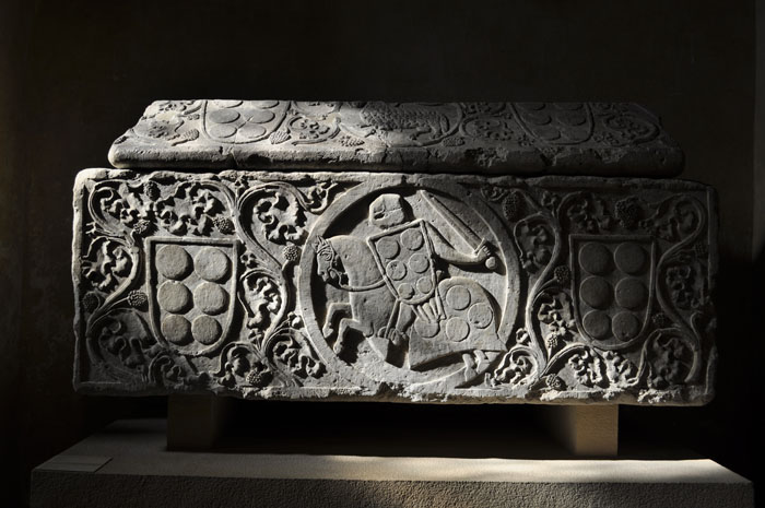 Tomb in shadows decorated with scrolling vines and shields and central medallion enclosing helmeted soldier holding sword and decorated shield, and mounted on draped horse.