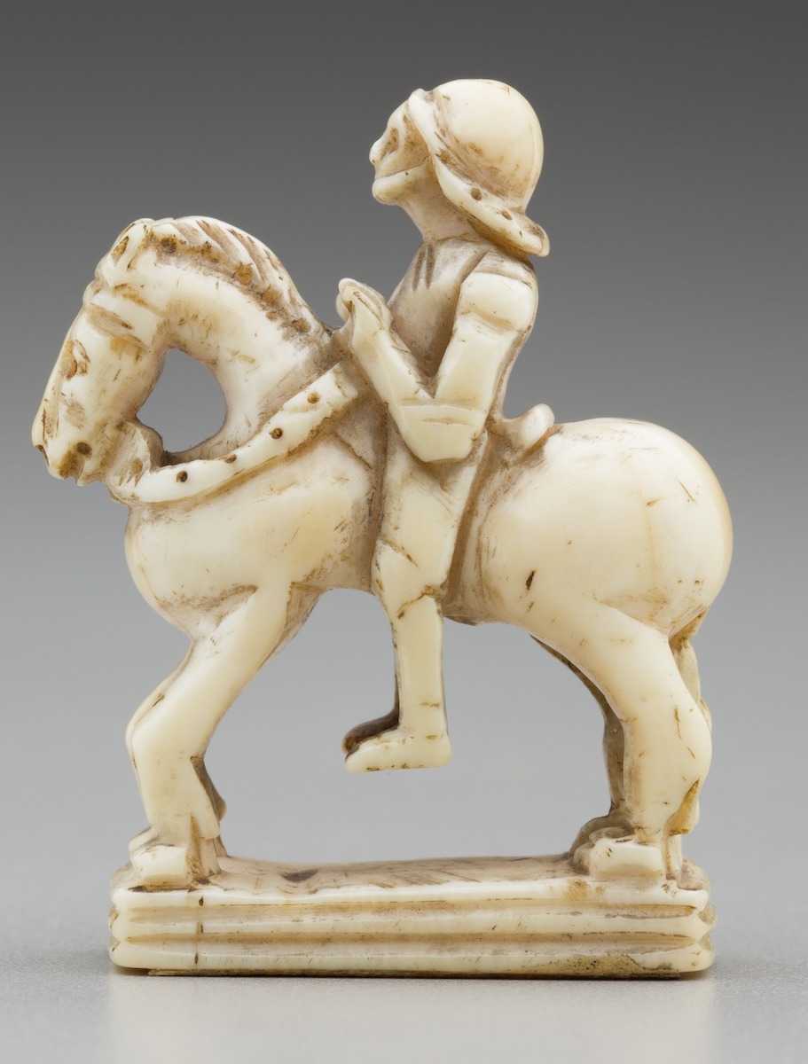 Carved ivory chess piece in the form of a knight seated astride a horse, wearing helmet and holdings the horse's reins, all on rectangular base.