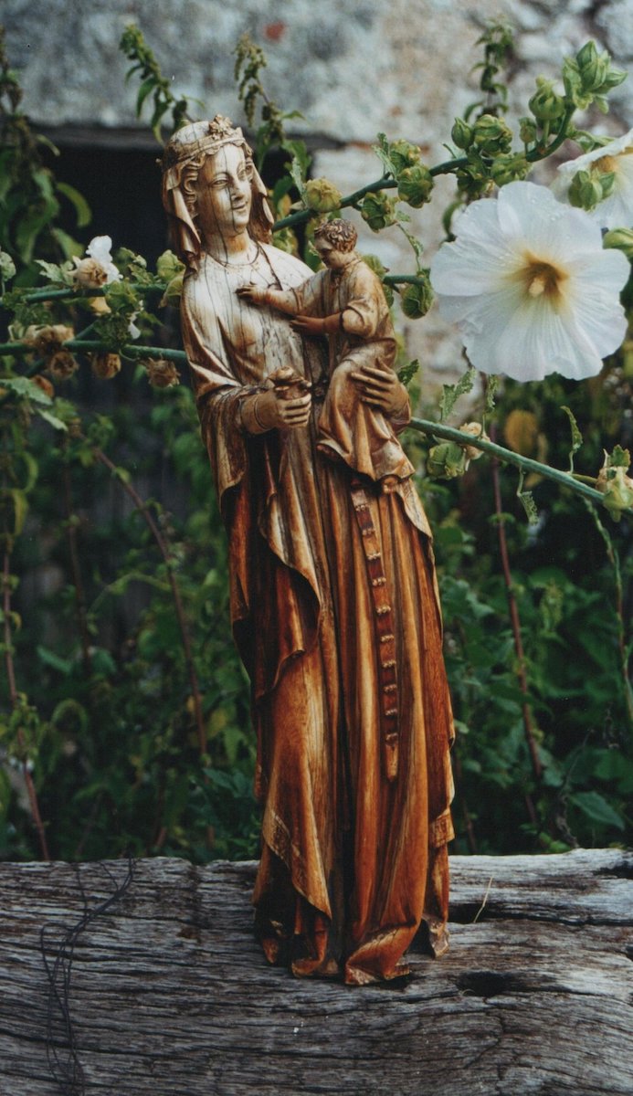 Carved ivory statuette of a veiled woman, wearing long belted garment, holding a male child in her left arm and a bud of a flower in her other hand. The statuette is on a wooden base surrounded by flowers. Some surface cracks and yellowing are present on the statue.