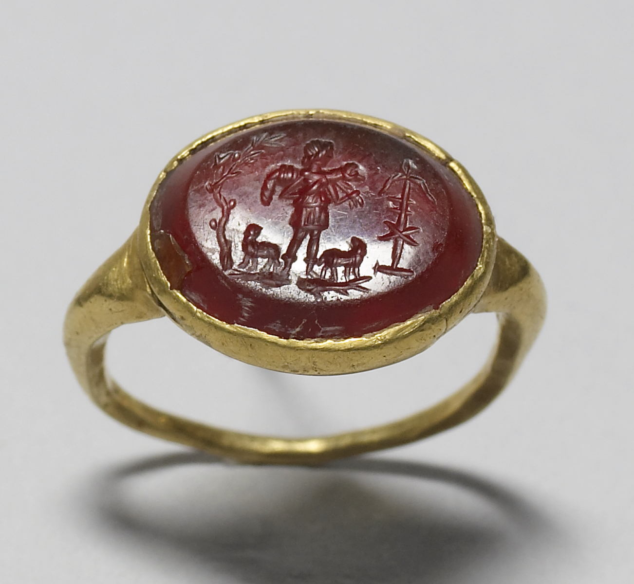 Gold jewelry ring set with oval red gem carved with scene of a young man, wearing tunic, standing on fish, holding sheep over shoulders and flanked by two sheep and a tree with dove and anchor with monogram.