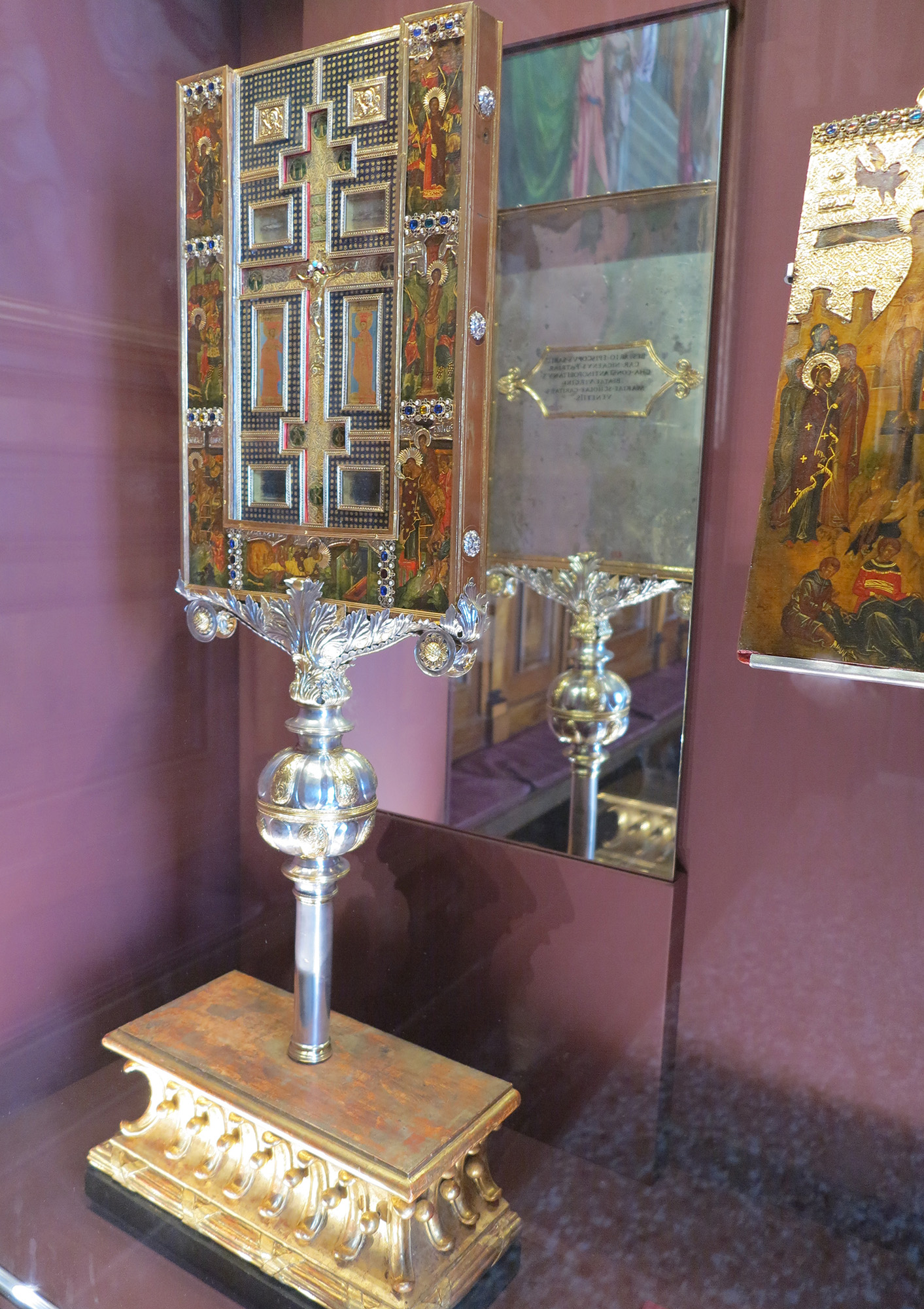 A painted reliquary with a metalwork cross, on a stand of metal on a wooden pedestal, open with its cover at the right, all before a mirror showing the reverse.