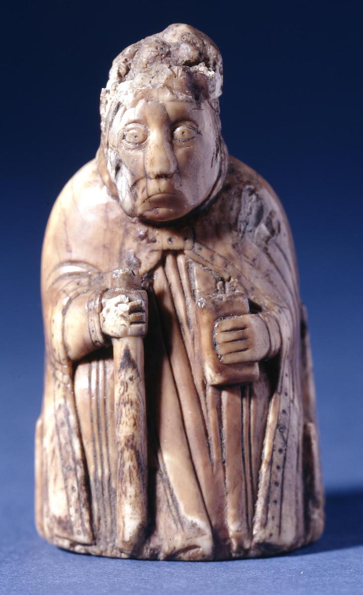 Carved ivory chess piece of mitered bishop (part of miter missing), wearing vestments, holding a crozier (head missing) and a book.