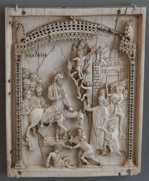 Carved rectangular ivory panel depicting a bearded man wearing a cross in halo and seated sideways on a saddled ass, followed by a group of bearded men, and the foreground two male figures are spreading a garment, and a youthful male seated on the ground looking at his foot. At the right, figures emerge from a city gate and two youthful figures climb a tree. The scene beneath an openwork canopy and Greek inscription in background.