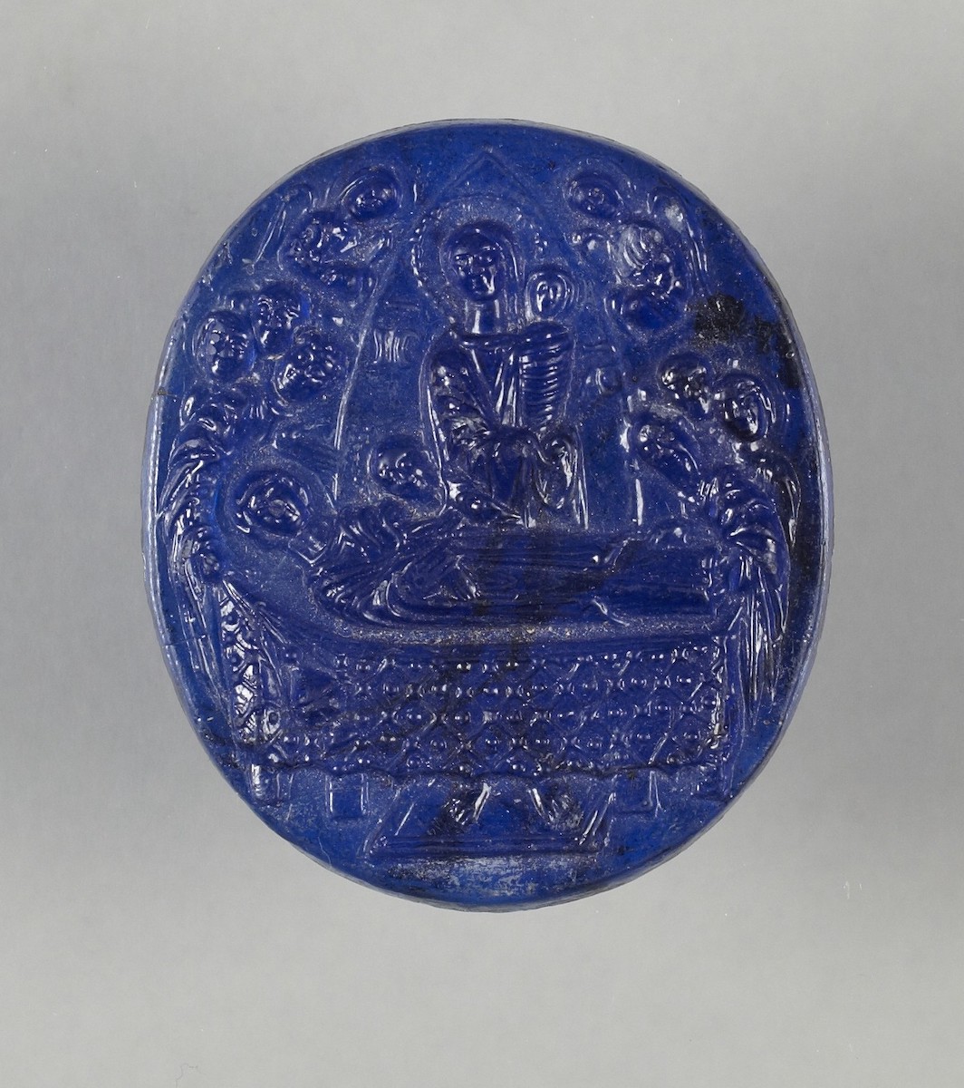 Blue oval cameo carved with man, with halo around his head, inscription in Greek on either side of him, standing in triangular opening, holding swaddled infant behind bed on which is veiled woman, with halo around her head, reclining in profile atop decorated blanket; feet of man visible on platform beneath bed. Above, busts of two angels and on either side heads of seven men, all with the suggestion of haloes around their heads.