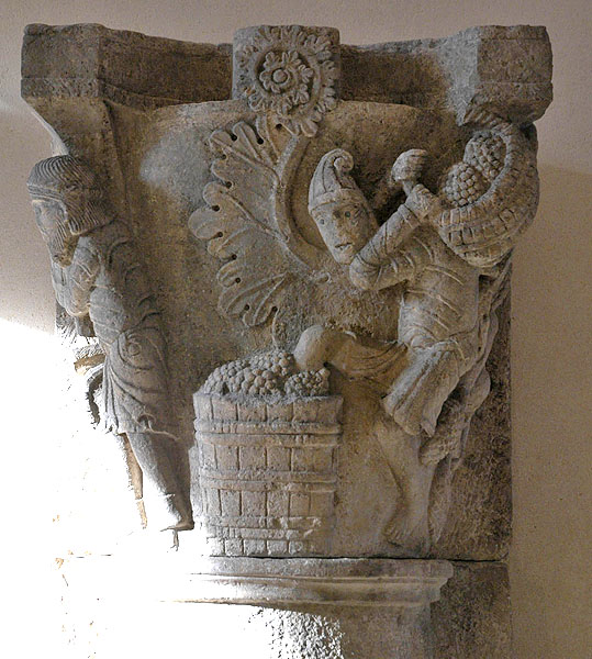 Detail of stone capital containing man carrying a basket of grapes on his shoulders and stepping into a vat of grapes with his left leg. 