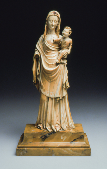 An ivory statuette of a veiled woman, draped with cloth over her long dress, holding in her left arm a male infant. The woman and child are both missing their right hands. The statuette is affixed to a stepped stone base against a studio background.