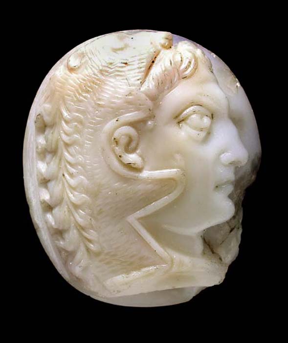 A carved oval, pink and white agate cameo depicting the profile head of a man, looking to right, wearing a lion’s skin over his head. Small chip on lower right portion of the cameo. Black background. 