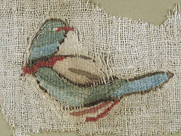 A decorated textile fragment depicting a bluish-green parrot standing in left facing profile with its head turned over its wing, and orange beak, neck feathers, and claws, all against white textile ground with frayed edges.
