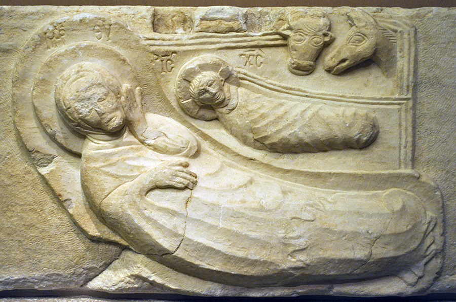 Detail of a relief sculpture of a woman lying next to a swaddled child with two animal heads above.