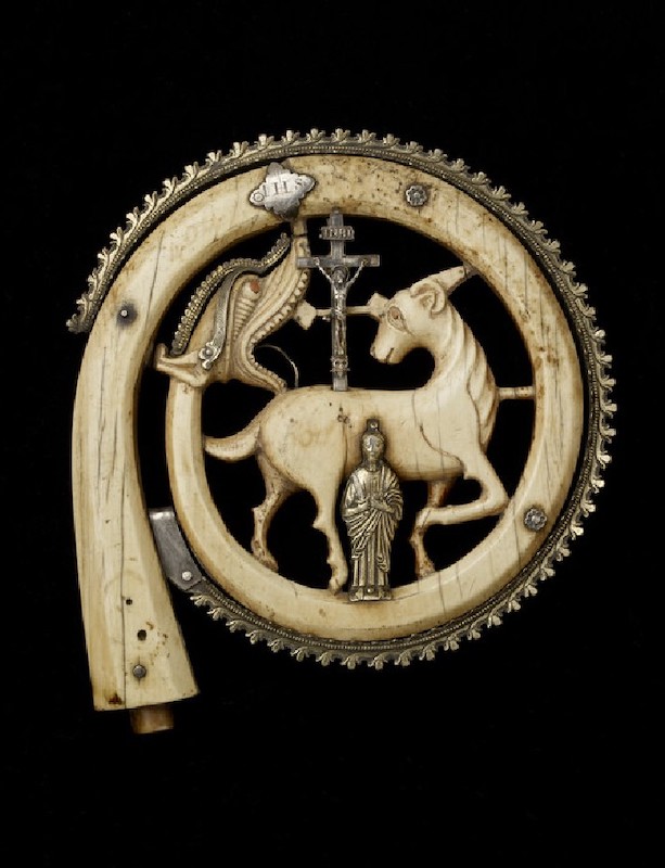 Spiral of carved ivory terminating in the head of a dragon with open jaws and enclosing a lamb in profile, looking back over cross. Silver figure, crucifix, IHS sign, figure, and foliate ornament on outer edge.
