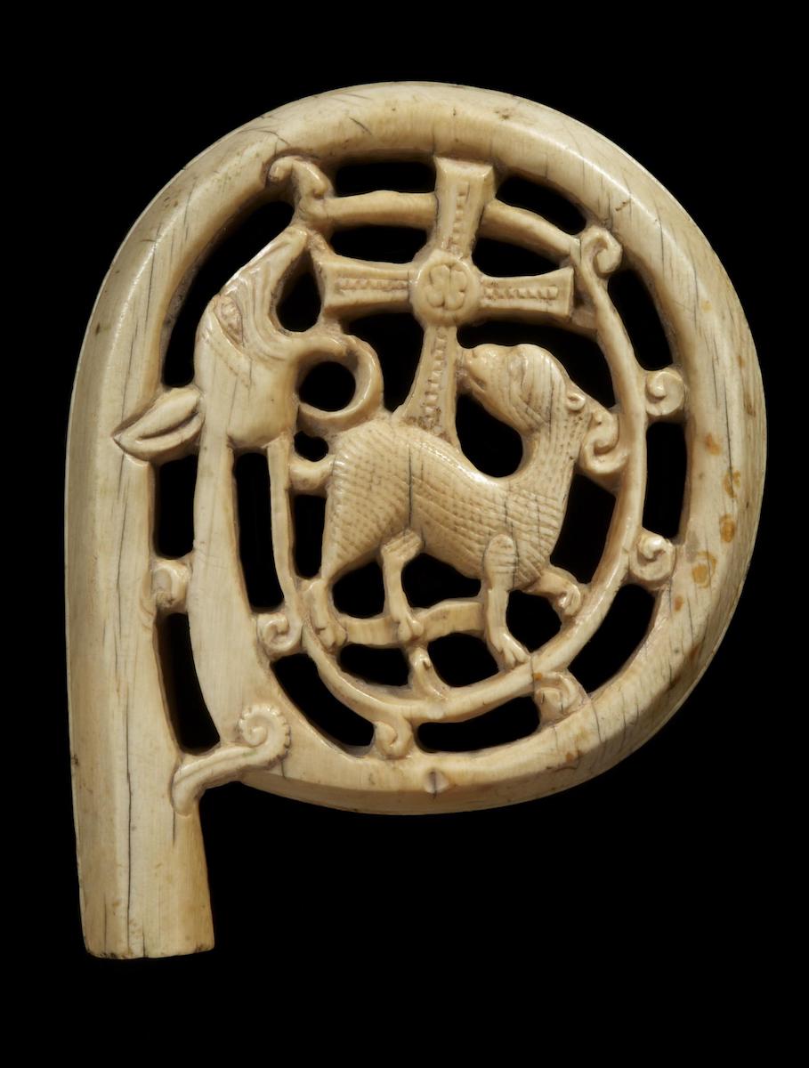 Head of an ivory crozier carved with scrolling volute enclosing a lamb, looking back to a cross, in jaws of the head of a dragon or serpent. Against black background.