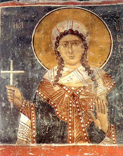 A fresco detail depicting the half figure of a woman wearing a white veil over her brown hair and a twisted scarf over a decorated dress in red, black, and white. Behind her head is a gold halo and she is holding a white cross and raising her left hand with her palm facing forward. Inscriptions of Greek letters surrounding the halo. Some paint surface wear.
