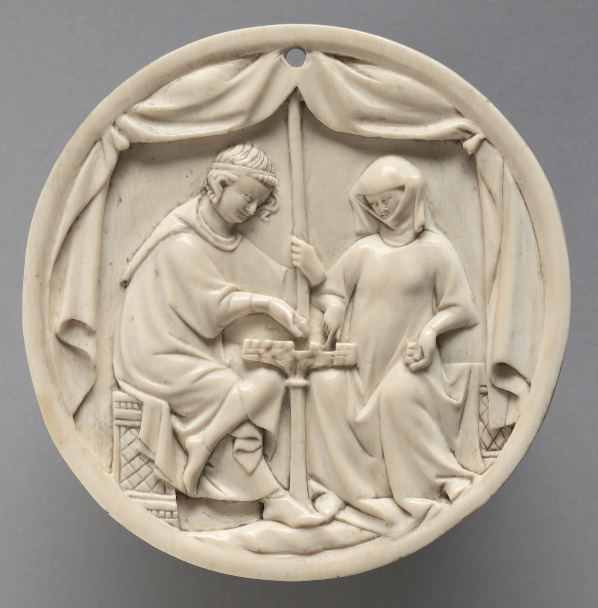 Round ivory plaque carved with a scene of a male and female seated in a tent around a chess board. At the top of the scene is a small drill hole.