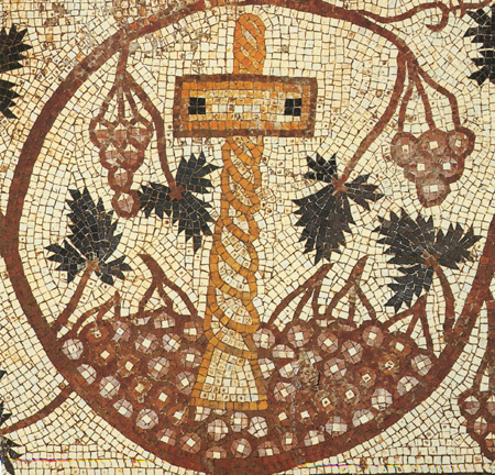 Mosaic detail of a medallion containing a wine press crushing grapes on the ground and surrounded by leafy grapevine with clusters of grapes. White background.
