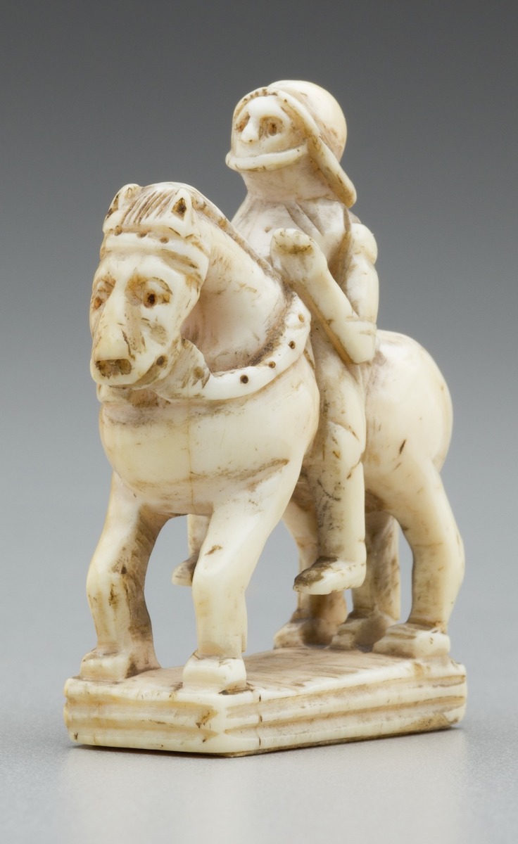 Carved ivory chess piece in the form of a knight seated astride a horse, wearing helmet and holdings the horse's reins, all on rectangular base.
