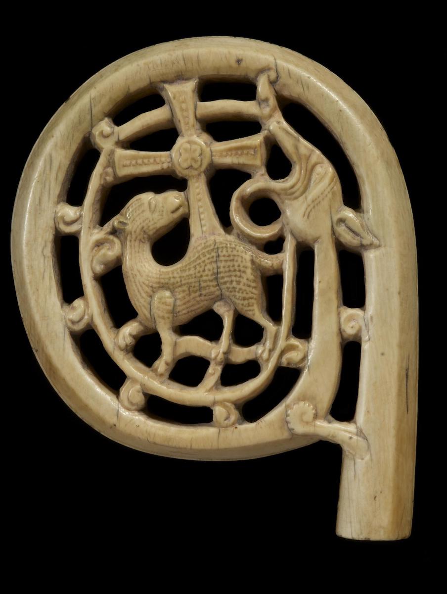 Head of an ivory crozier carved with scrolling volute enclosing a lamb, looking back to a cross, in jaws of the head of a dragon or serpent. Against black background.