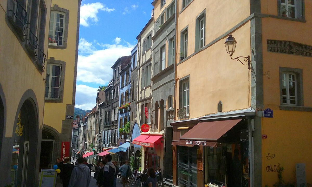 People walk along a street lined with buildings with colorful awnings. At the corner is a yellow building supporting a streetlamp. Embedded in one wall of the yellow building is a stone lintel carved with a kneeling man washing the feet of another man, the two men surrounded by eleven other men.