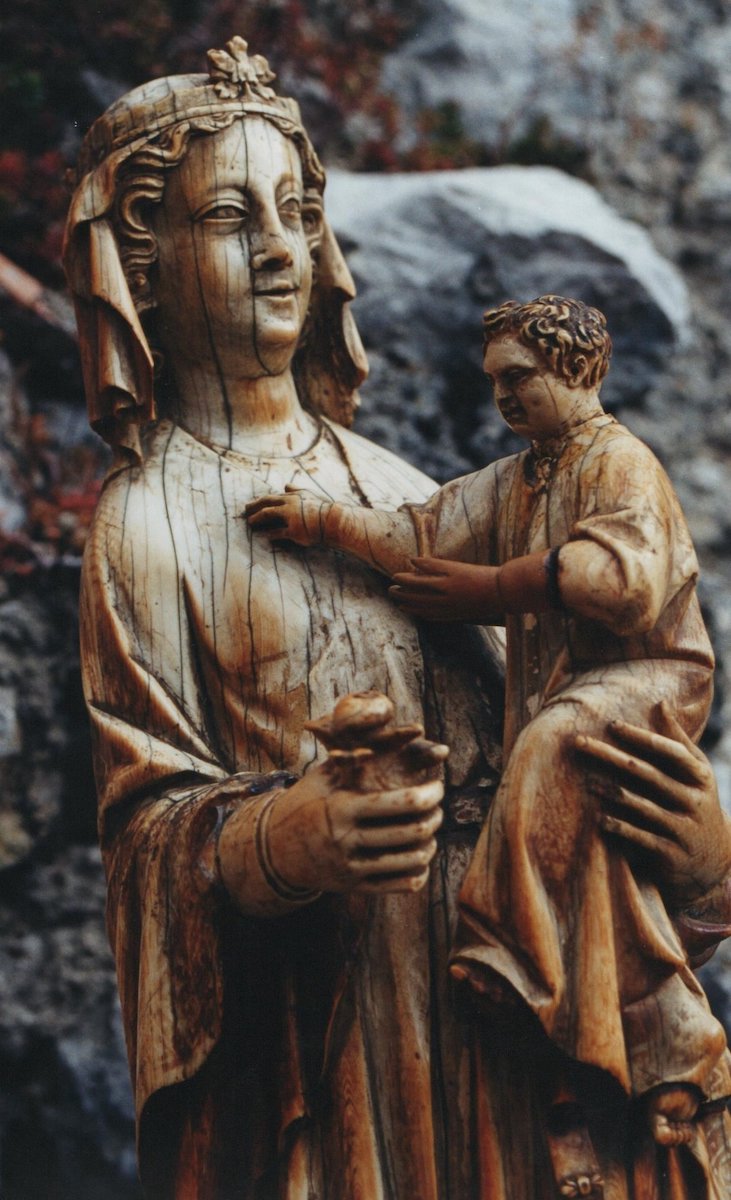 A detail of a carved ivory statuette of a veiled woman, her head and bust only, holding a male child in her left arm and a bud of a flower in the other hand. Some surface cracks and yellowing are present on the statue.