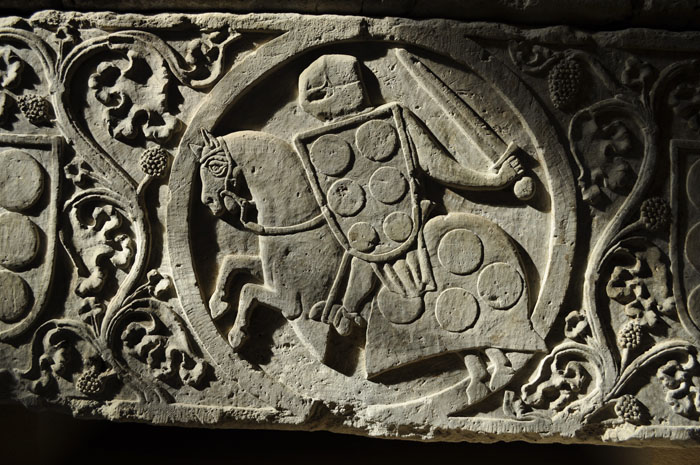 Sculptural detail of tomb showing medallion enclosing helmeted soldier holding sword and decorated shield, mounted on draped horse, and surrounded by scrolling vines and shields.