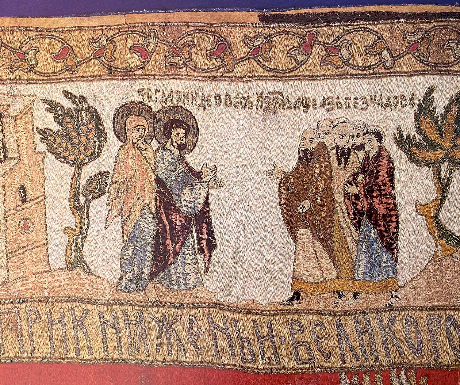 In a detail of an embroidered veil, a woman and a man with his hands held out before him face a group of five men, all between two trees.