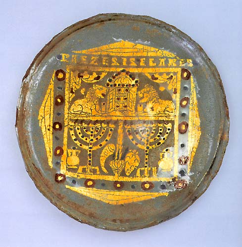 A glass disk with a design in gold, featuring a tabernacle, two lions, two menorahs, and other religious implements.