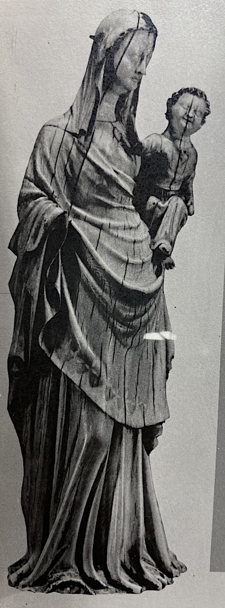 From the side, an ivory statuette of a veiled woman, draped with cloth over her long dress, holding in her left arm a male infant. The woman and child are both missing their right hands. 