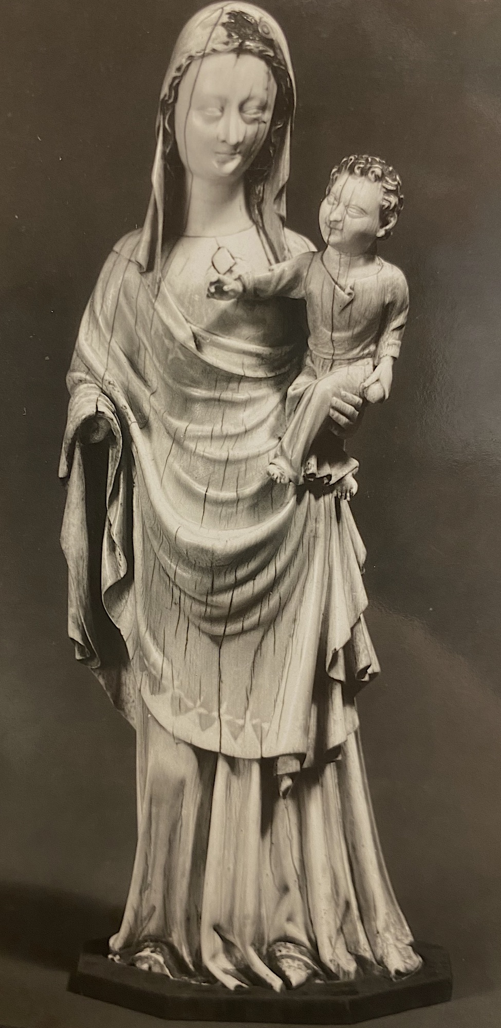 An ivory statuette of a veiled woman, draped with cloth over her long dress, holding in her left arm a male infant. The woman and child are both missing their right hands. The statuette is affixed to a stepped stone base against a studio background.