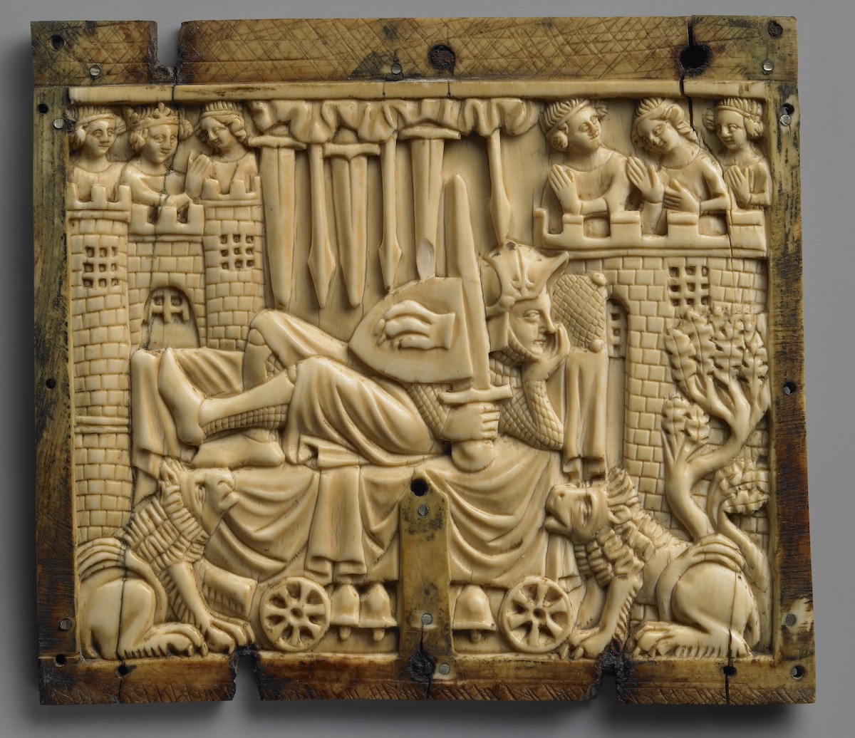 A squarish ivory panel carved with the scene of a man, wearing a helmet and full armor, reclining on a wheeled bed with bells, beneath swords and spears falling from sky, and between two lions and castle towers with female figures on each top. Border with yellowing, hatching, and some cracks with loss.