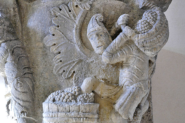 Detail of stone capital containing man carrying a basket of grapes on his shoulders and stepping into a vat of grapes with his left leg. 
