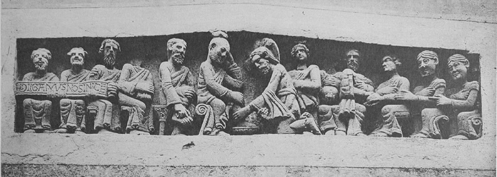 In a carved lintel, a kneeling man washes the feet of another man, the two men surrounded by eleven other men.