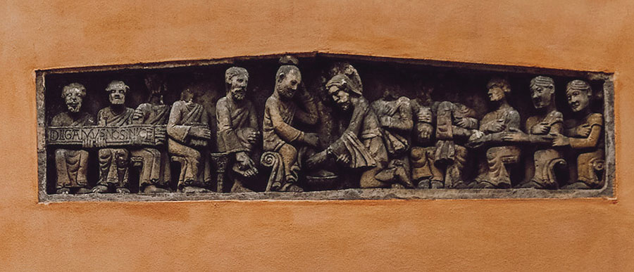 In a carved lintel, a kneeling man washes the feet of another man, the two men surrounded by eleven other men.