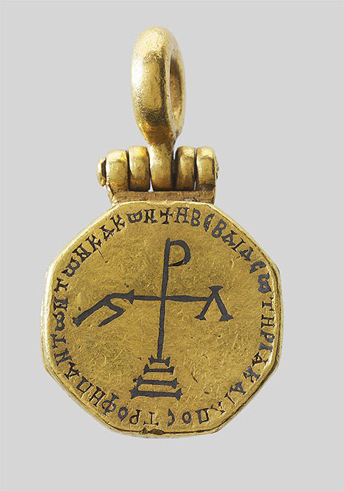 The bottom cover of a gold reliquary pendant with an inscription in Greek surrounding a cross with Greek numerals at the end of its arms standing on a base and three steps.